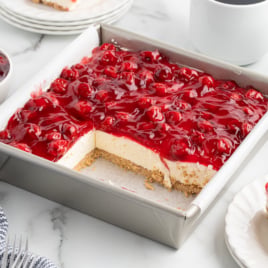 A square pan of No Bake Cherry Cheesecake with a graham cracker crust sits on a marble surface. One section has been cut and served on a plate nearby. Bowls of cherries, forks, and cups are also visible.