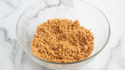 A glass bowl filled with a crumbly mixture of crushed graham crackers and sugar sits on a white marble surface, ready to become the perfect crust for a delicious No Bake Cherry Cheesecake.