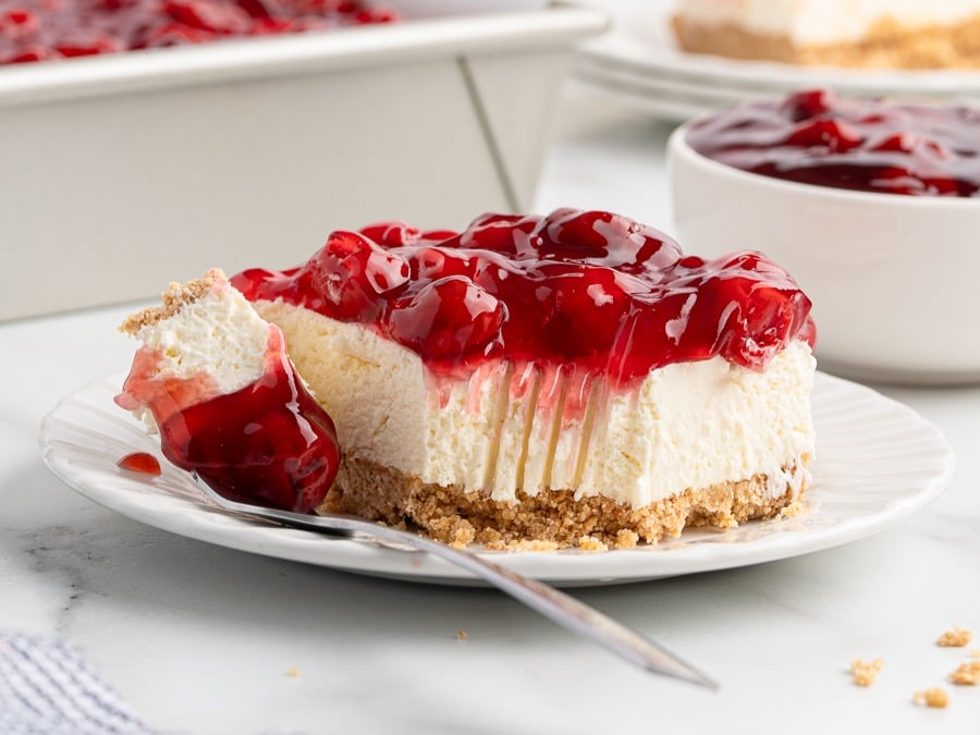 A slice of No Bake Cherry Cheesecake with a graham cracker crust sits on a white plate. A bite reveals the creamy filling, while a spoon rests nearby and a bowl of cherry topping is in the background.