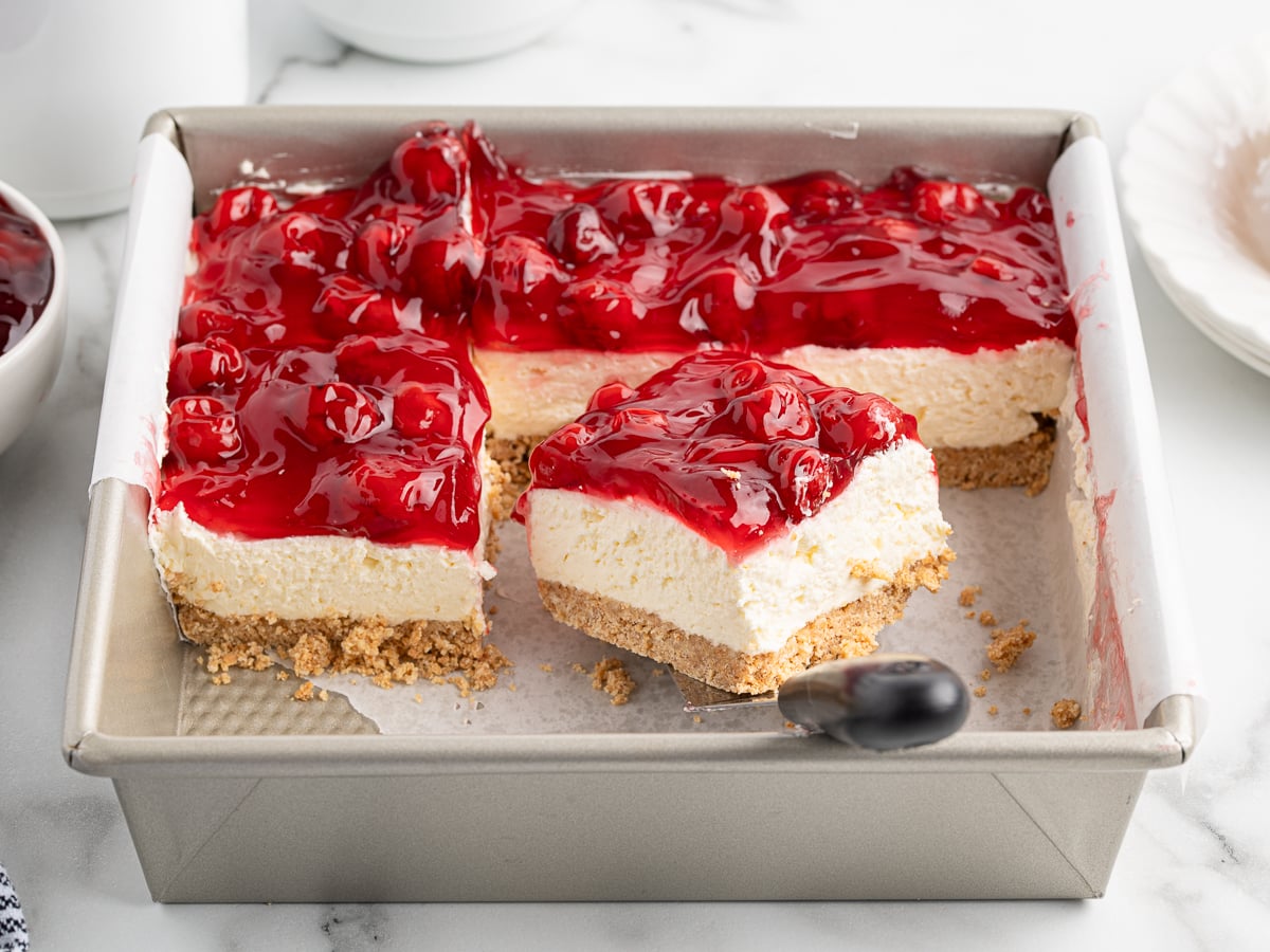 A square baking pan holds a No Bake Cherry Cheesecake topped with cherry sauce. Two large slices are cut, and one slice is being lifted out with a spatula, showing the graham cracker crust and creamy filling.