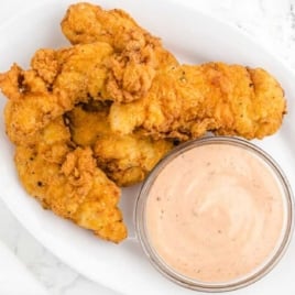 Oval plate with crispy chicken fingers served alongside a bowl of creamy dipping sauce on a white marble surface.