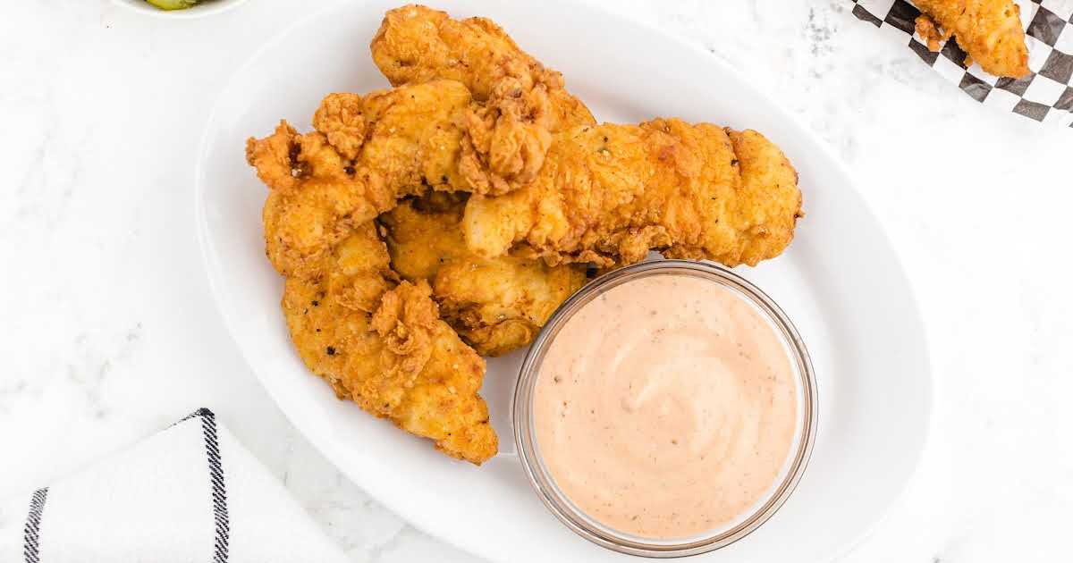 Oval plate with crispy chicken fingers served alongside a bowl of creamy dipping sauce on a white marble surface.