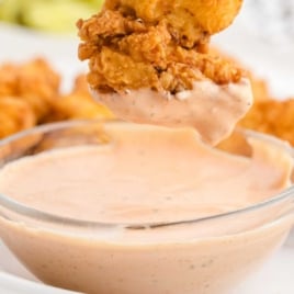 Fried chicken tender dipped into creamy orange dipping sauce in a glass bowl, with blurred chicken fingers in the background.