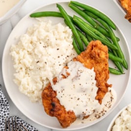 a plate of chicken fried chicken topped with gravy and served with mashed potatoes and green beans