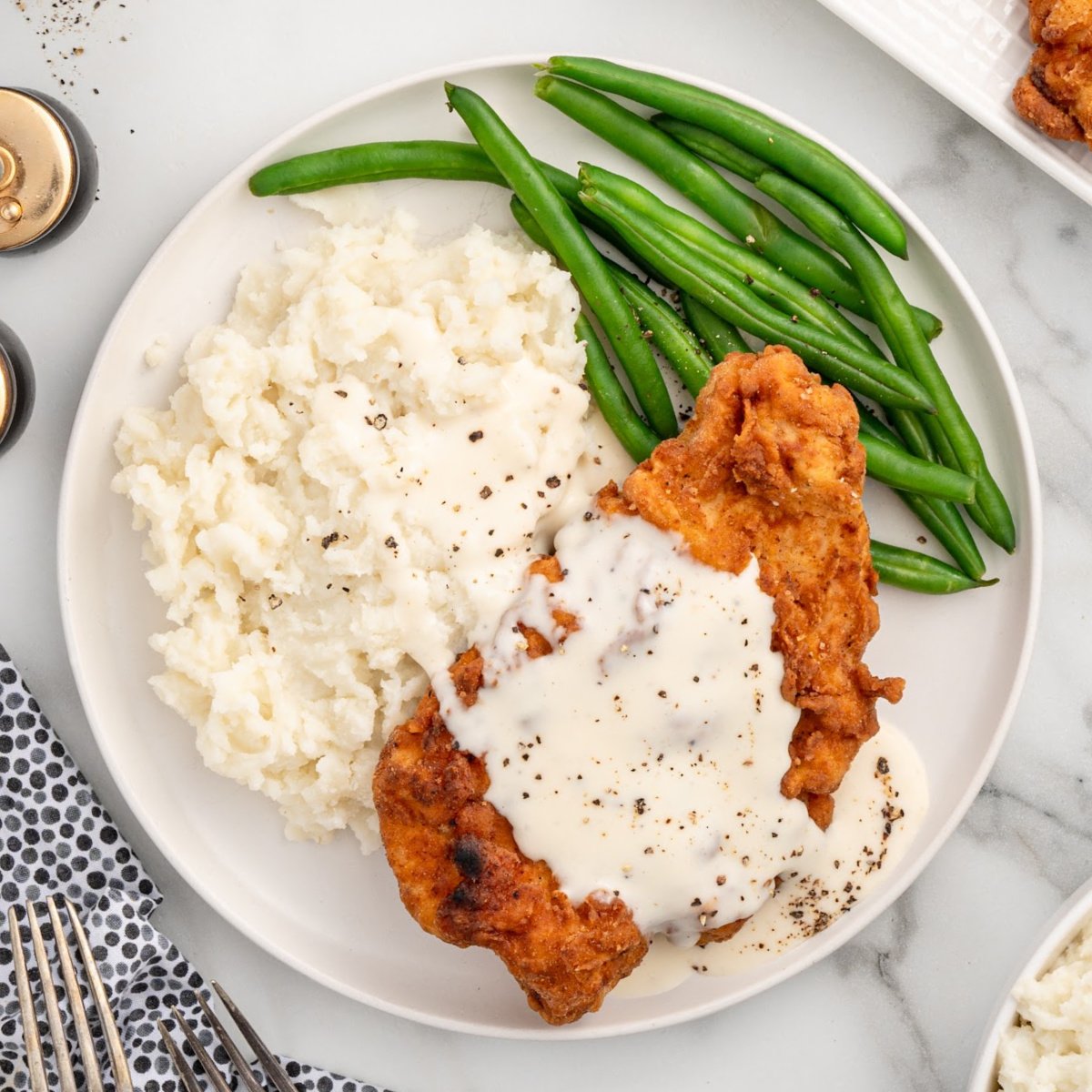 a plate of chicken fried chicken topped with gravy and served with mashed potatoes and green beans