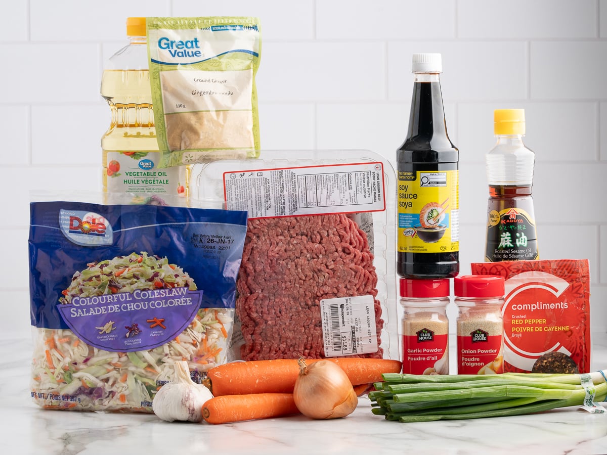 Assorted ingredients for an Egg Roll in a Bowl recipe are arranged on a counter&mdash;ground beef, coleslaw mix, carrots, onions, green onions, vegetable oil, soy sauce, sesame oil, garlic powder, red pepper, and ground ginger&mdash;against a white tile background.