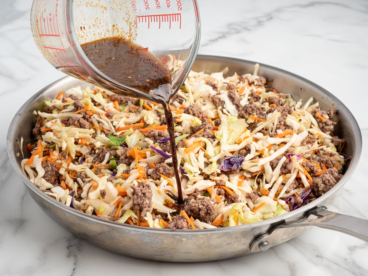 A glass measuring cup pours a dark sauce over a skillet filled with cooked ground beef, shredded cabbage, carrots, and veggies&mdash;perfect for an easy Egg Roll in a Bowl Recipe on a marble countertop.