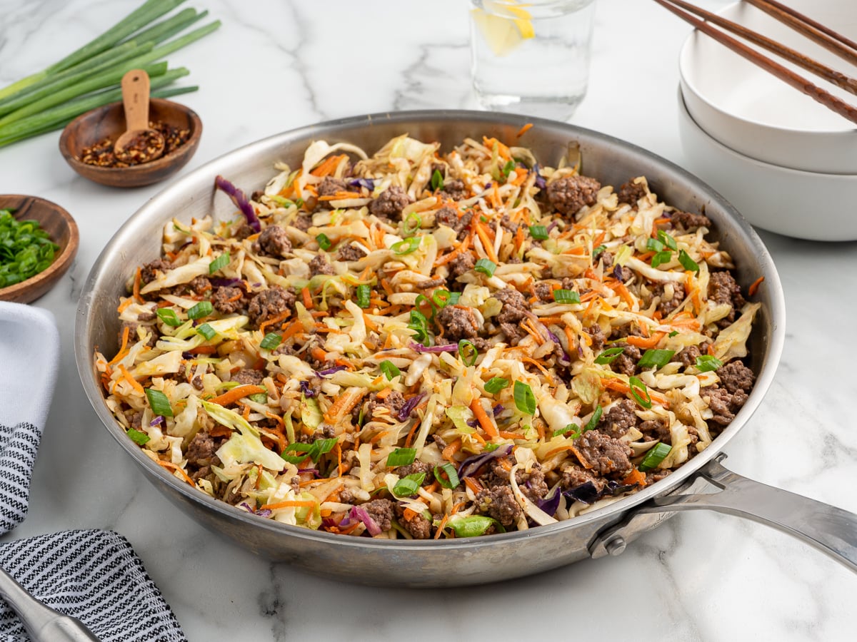 A stainless steel skillet filled with a savory Egg Roll in a Bowl stir-fry&mdash;ground beef, shredded cabbage, carrots, and green onions&mdash;sits on a marble countertop with bowls, chopsticks, and seasonings nearby.