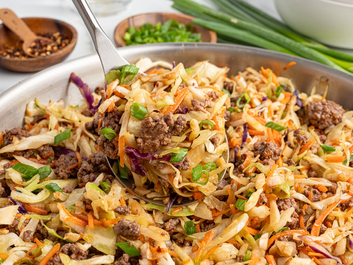 A close-up of a skillet filled with cooked ground beef, shredded cabbage, carrots, and green onions&mdash;the classic Egg Roll in a Bowl Recipe. A spoon lifts a portion, with bowls of sliced green onions and red pepper flakes in the background.