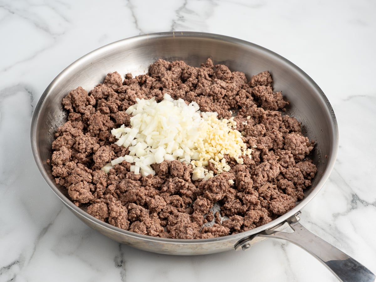 Ground beef is cooking in a stainless steel skillet on a marble countertop, with chopped raw onions and minced garlic added on top but not yet stirred in&mdash;perfect beginnings for an Egg Roll in a Bowl recipe.