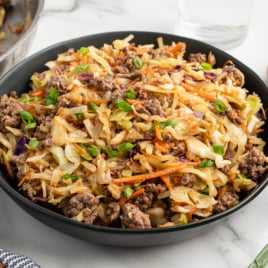 A black bowl filled with Egg Roll in a Bowlโground beef, shredded cabbage, carrots, and green onionsโsits on a white surface, with a small bowl of chopped green onions and seasonings nearby. Try this Egg Roll in a Bowl Recipe for a quick meal!.