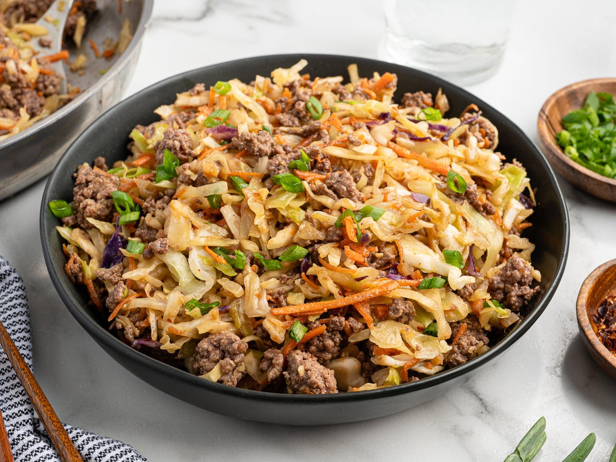 A black bowl filled with Egg Roll in a Bowlโground beef, shredded cabbage, carrots, and green onionsโsits on a white surface, with a small bowl of chopped green onions and seasonings nearby. Try this Egg Roll in a Bowl Recipe for a quick meal!.