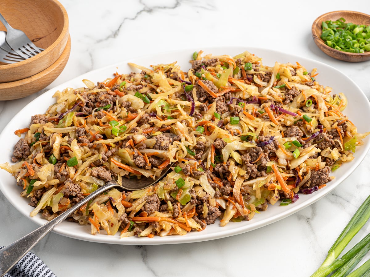 A white platter filled with Egg Roll in a Bowl&mdash;ground beef, shredded cabbage, carrots, and green onions stir-fried together. A metal serving spoon rests on the platter, with a small bowl of sliced green onions nearby on the marble surface.