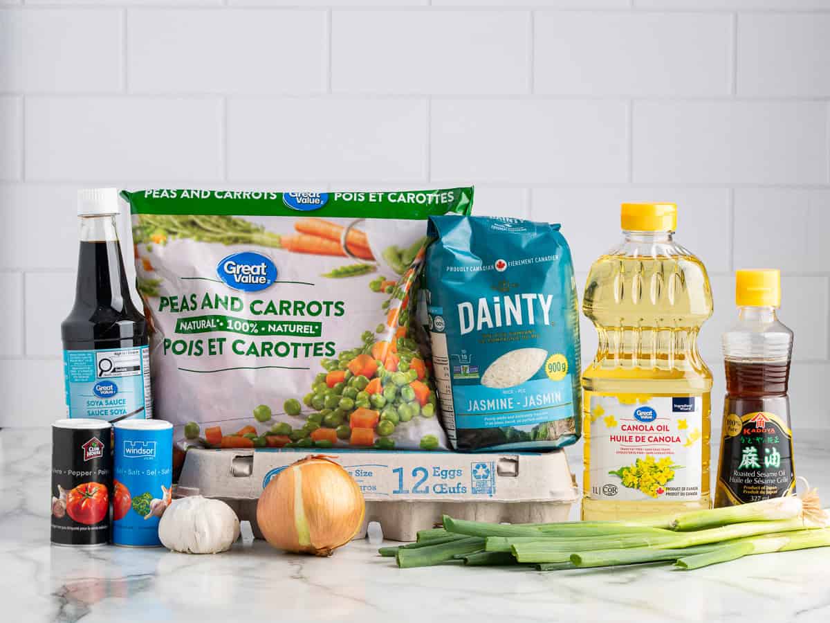 A selection of Better Than Take-Out Fried Rice ingredients on a counter, including jasmine rice, peas and carrots, soy sauce, eggs, canola oil, sesame oil, green onions, garlic, onion, salt, and black pepper.