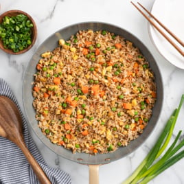 A skillet filled with Better Than Take-Out Fried Rice, featuring peas, carrots, and green onions. Surrounding the skillet are bowls of sliced green onions, pepper, salt, a soy sauce dish, chopsticks, a wooden spoon, and a kitchen towel.