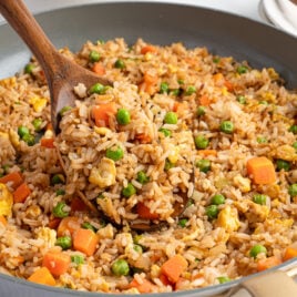 A wooden spoon scoops Better Than Take-Out Fried Rice with peas, diced carrots, and scrambled eggs from a large skillet. The rice is evenly mixed and cooked, with visible chunks of vegetables and egg.