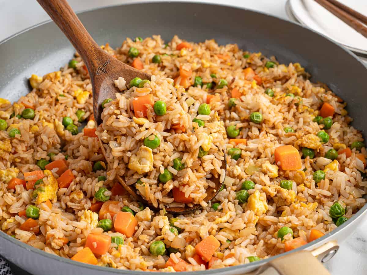 A wooden spoon scoops Better Than Take-Out Fried Rice with peas, diced carrots, and scrambled eggs from a large skillet. The rice is evenly mixed and cooked, with visible chunks of vegetables and egg.