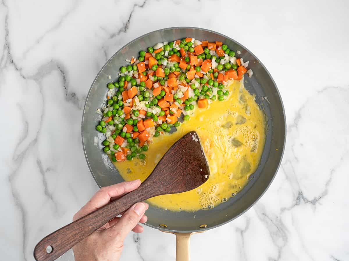A hand stirs scrambled eggs in a skillet with diced carrots, peas, and chopped onions on a white marble surfaceโthe perfect start to a Better Than Take-Out Fried Rice Recipe.