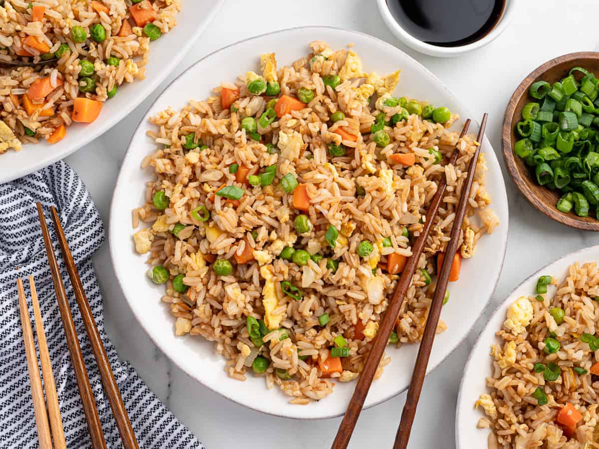 A plate of Better Than Take-Out Fried Rice with peas, carrots, and scrambled eggs, garnished with green onions. Chopsticks rest on the plate, with bowls of soy sauce and extra green onions nearby.