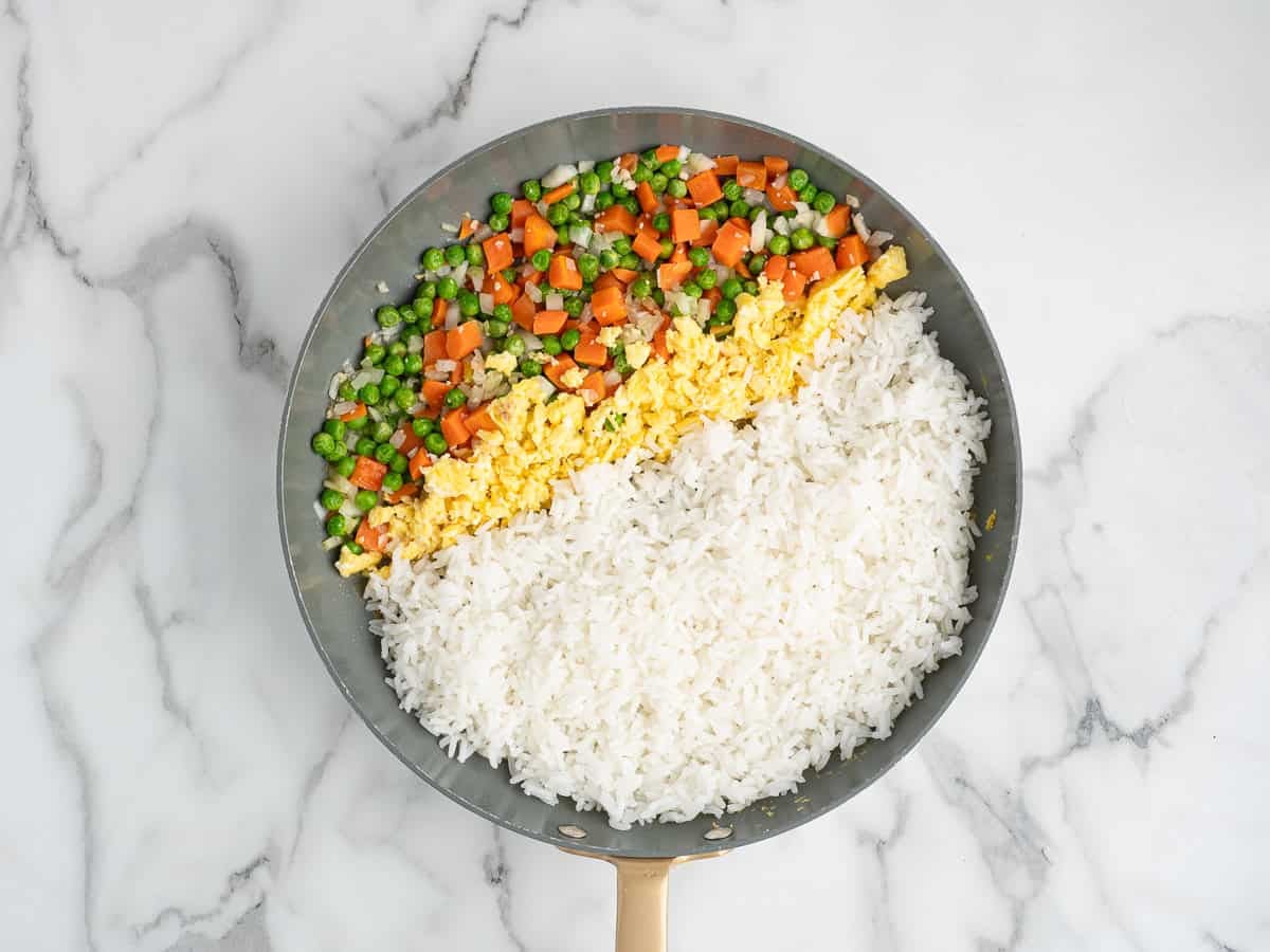 A frying pan on a marble surface contains separated sections of cooked white rice, scrambled eggs, and a mix of chopped carrots and green beansโperfect for making a Better Than Take-Out Fried Rice recipe at home.