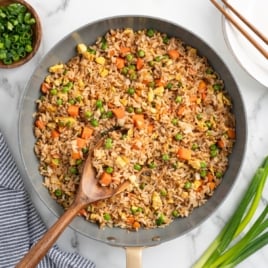 A skillet filled with Better Than Take-Out Fried Rice featuring peas, carrots, scrambled eggs, and green onions. A wooden spoon rests in the pan. Chopped and whole green onions are scattered nearby on a marble surface.