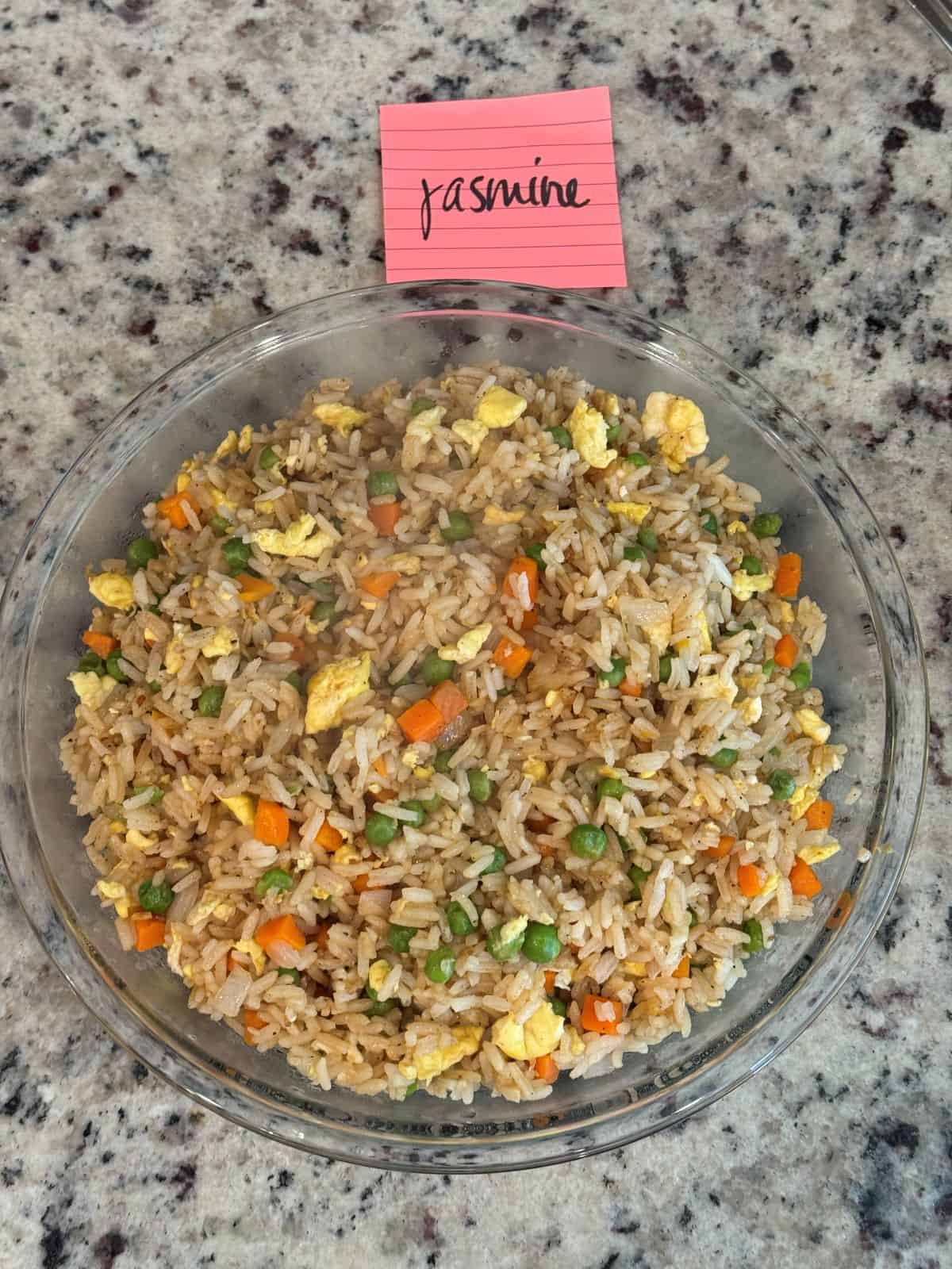 A glass bowl filled with Better Than Take-Out Fried Riceโmixed with peas, carrots, and scrambled eggsโsits on a speckled countertop. A pink sticky note labeled jasmine is placed above the bowl.