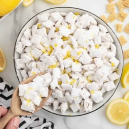 A bowl of Lemon Puppy Chow—a lemon-flavored Chex mix coated in powdered sugar and garnished with fresh lemon zest—sits at the center, surrounded by sliced lemons and scattered Chex cereal pieces. A hand holds a wooden spoon, ready to scoop this delicious Lemon Puppy Chow Recipe.