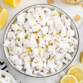 A bowl of Lemon Puppy Chow, a delightful lemon-flavored powdered sugar-coated cereal snack mix, garnished with lemon zest and surrounded by lemon slices and cereal pieces on a white surface—a refreshing twist on the classic Lemon Puppy Chow Recipe.