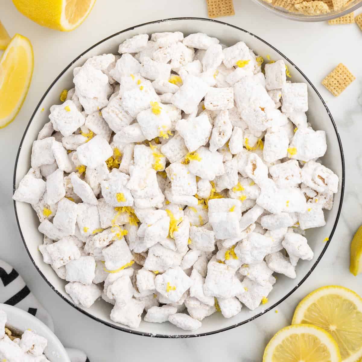 A bowl of Lemon Puppy Chow, a delightful lemon-flavored powdered sugar-coated cereal snack mix, garnished with lemon zest and surrounded by lemon slices and cereal pieces on a white surface—a refreshing twist on the classic Lemon Puppy Chow Recipe.