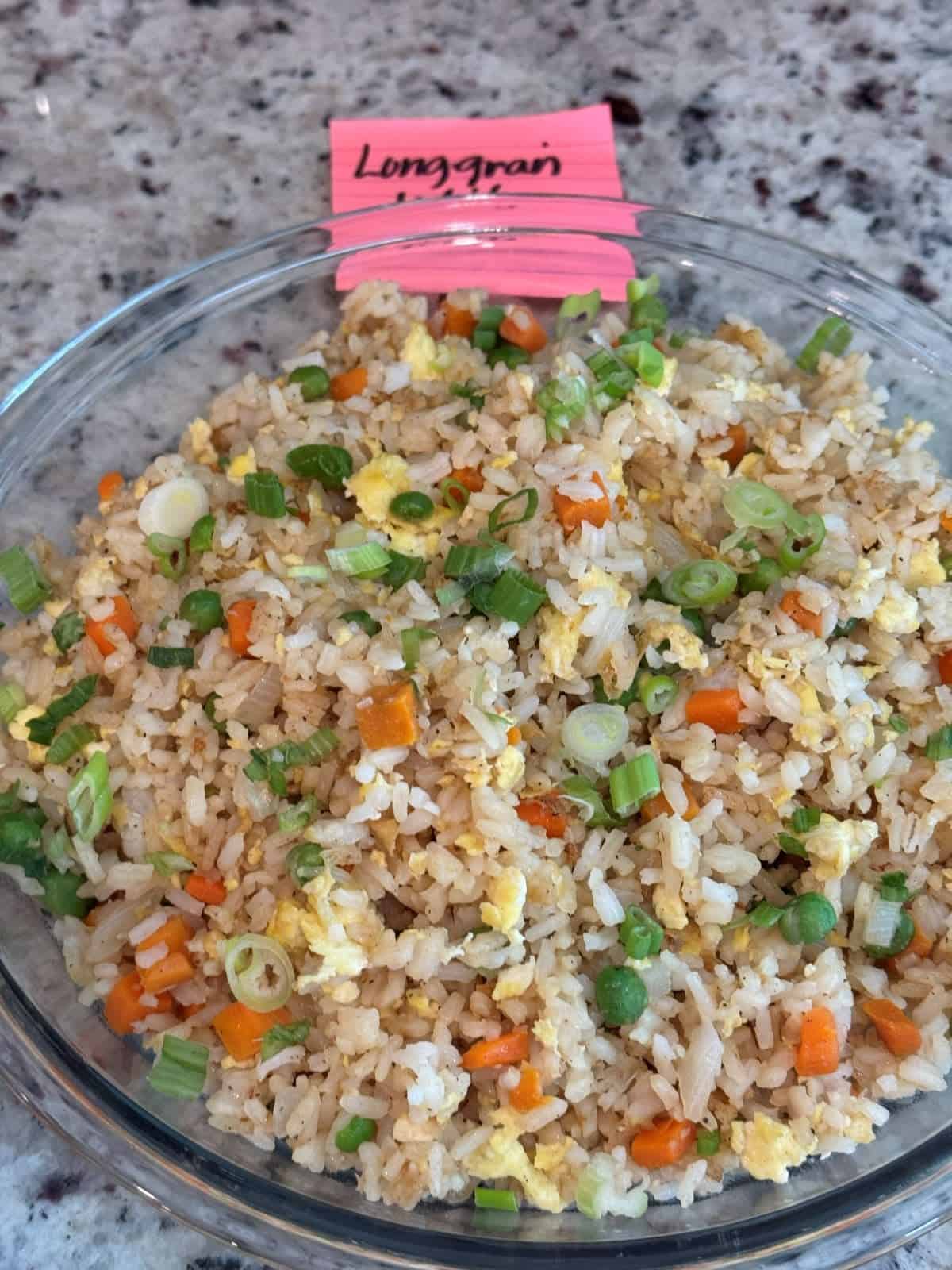 A glass bowl filled with Better Than Take-Out Fried Rice featuring peas, carrots, scrambled egg, and chopped green onions sits on a counter. A pink label behind the bowl reads Long grain.