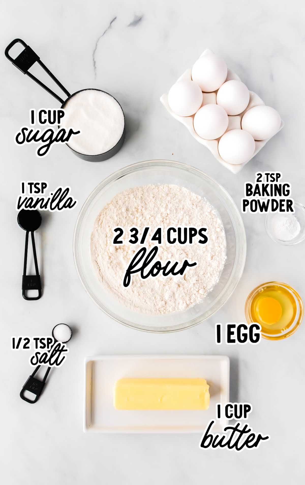 A flat lay of the ingredients for Easter cookies, including flour, sugar, vanilla, butter, eggs, and baking powder, labeled with measurements on a white marble surface.