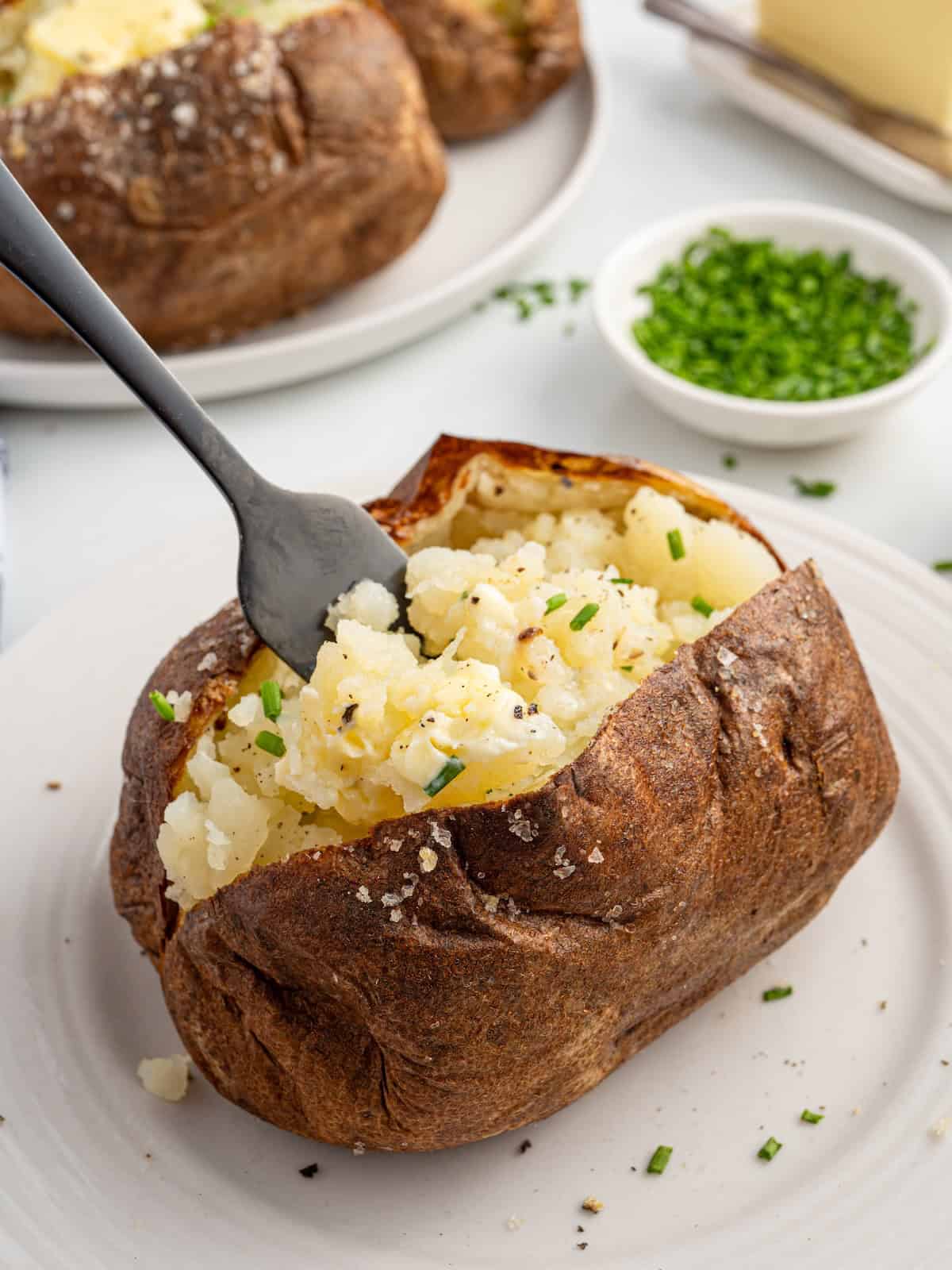 An Air Fryer Baked Potato split open on a white plate, topped with chopped chives and black pepper, with a fork fluffing the potato. A bowl of chives and a stick of butter are in the background.