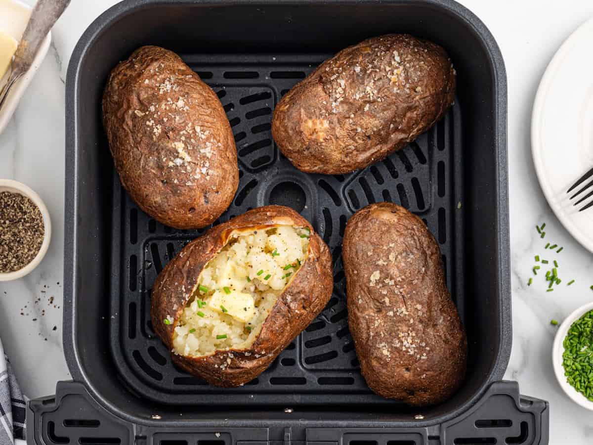Four Air Fryer Baked Potatoes, one split open and topped with chopped chives, sit in an air fryer basket. The potatoes are sprinkled with salt, and small dishes of pepper and chives are nearby.