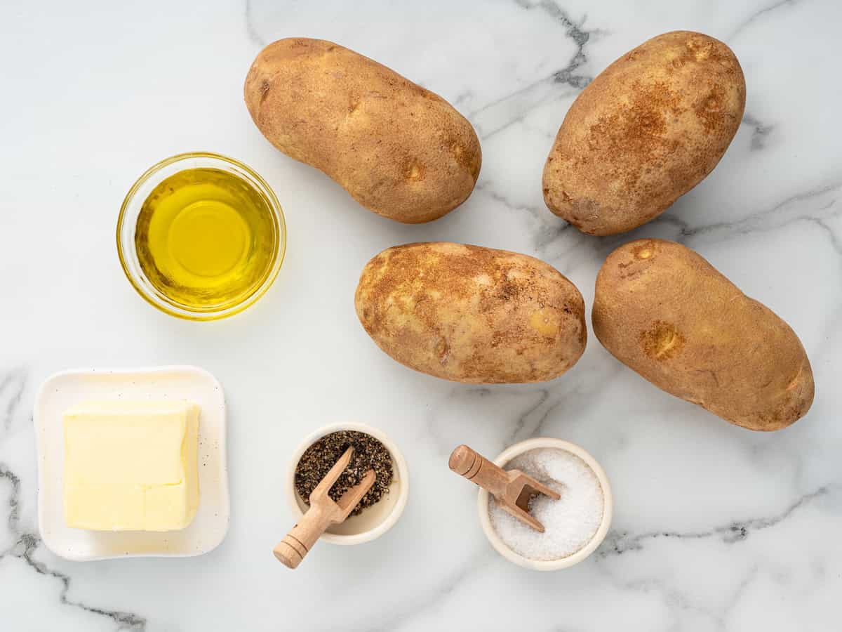 Four russet potatoes, a small bowl of olive oil, a plate with butter, and small bowls of salt and pepper are arranged on a white marble surface—perfect for making Air Fryer Baked Potato.