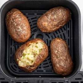 Four Air Fryer Baked Potatoes rest in an air fryer basket; one is split open, revealing a mashed, seasoned filling topped with chopped chives. Plates, utensils, and bowls of pepper and chives are nearby.