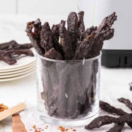 A clear glass filled with strips of dark Air Fryer Teriyaki Beef Jerky sits on a white surface, with plates, a spoon of red pepper flakes, and a checkered cloth in the background.