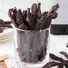 A clear glass filled with strips of dark Air Fryer Teriyaki Beef Jerky, with a few more pieces scattered around on a white surface.