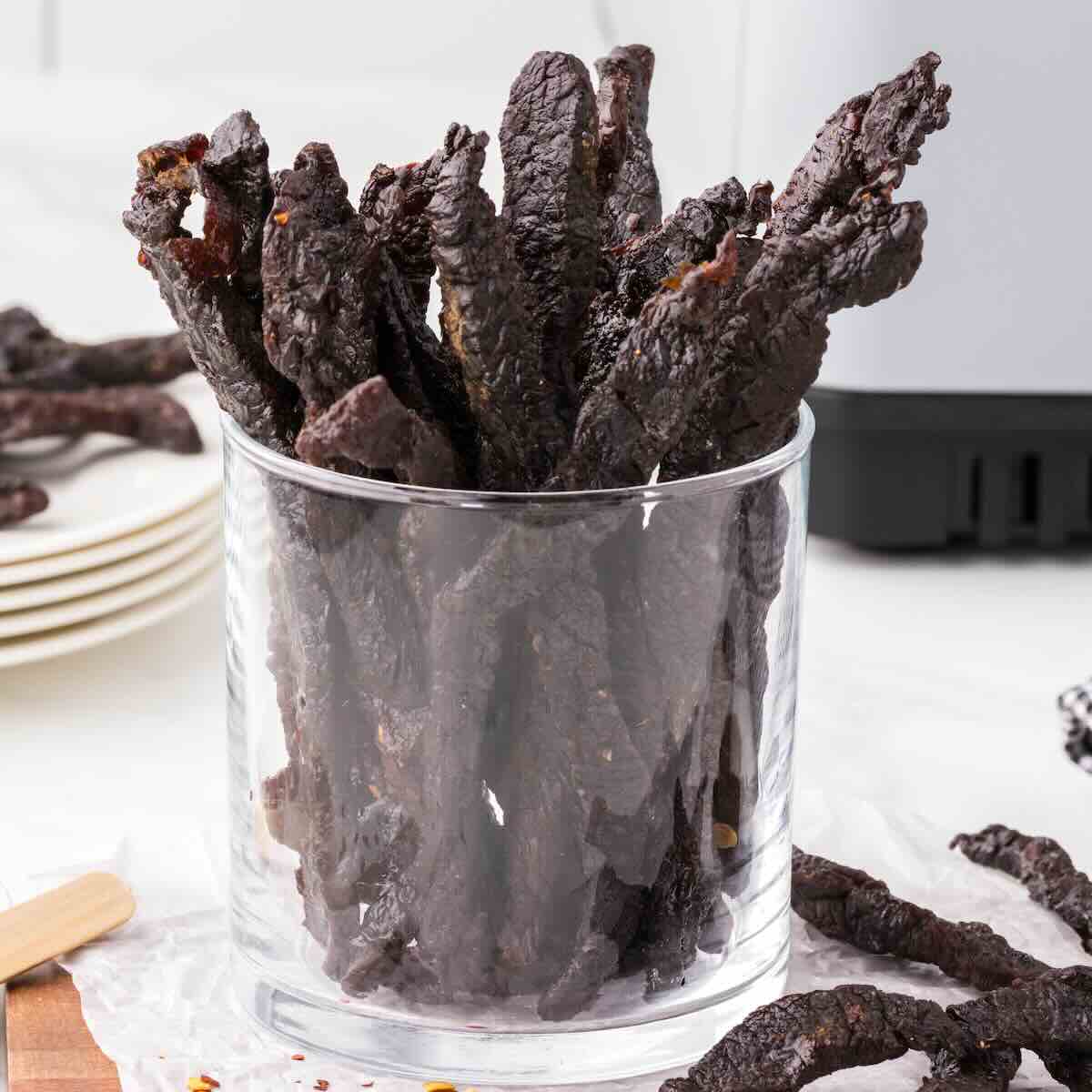 A clear glass filled with strips of dark Air Fryer Teriyaki Beef Jerky, with a few more pieces scattered around on a white surface.