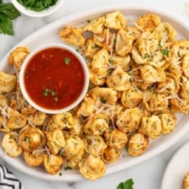 A platter of air fryer tortellini served with a bowl of marinara sauce, garnished with fresh parsley and shredded Parmesan, with additional ingredients in the background.