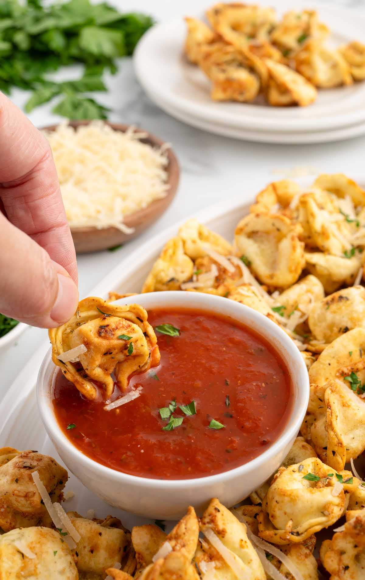 A person holding a tortellini dipped in marinara sauce over a plate filled with golden, crispy air fryer tortellini.