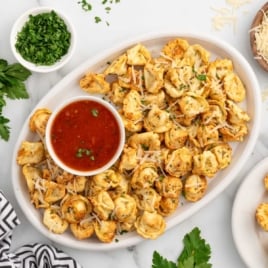 A round plate filled with golden air fryer tortellini garnished with parsley and Parmesan, accompanied by a bowl of marinara sauce, with additional parsley leaves on the side.