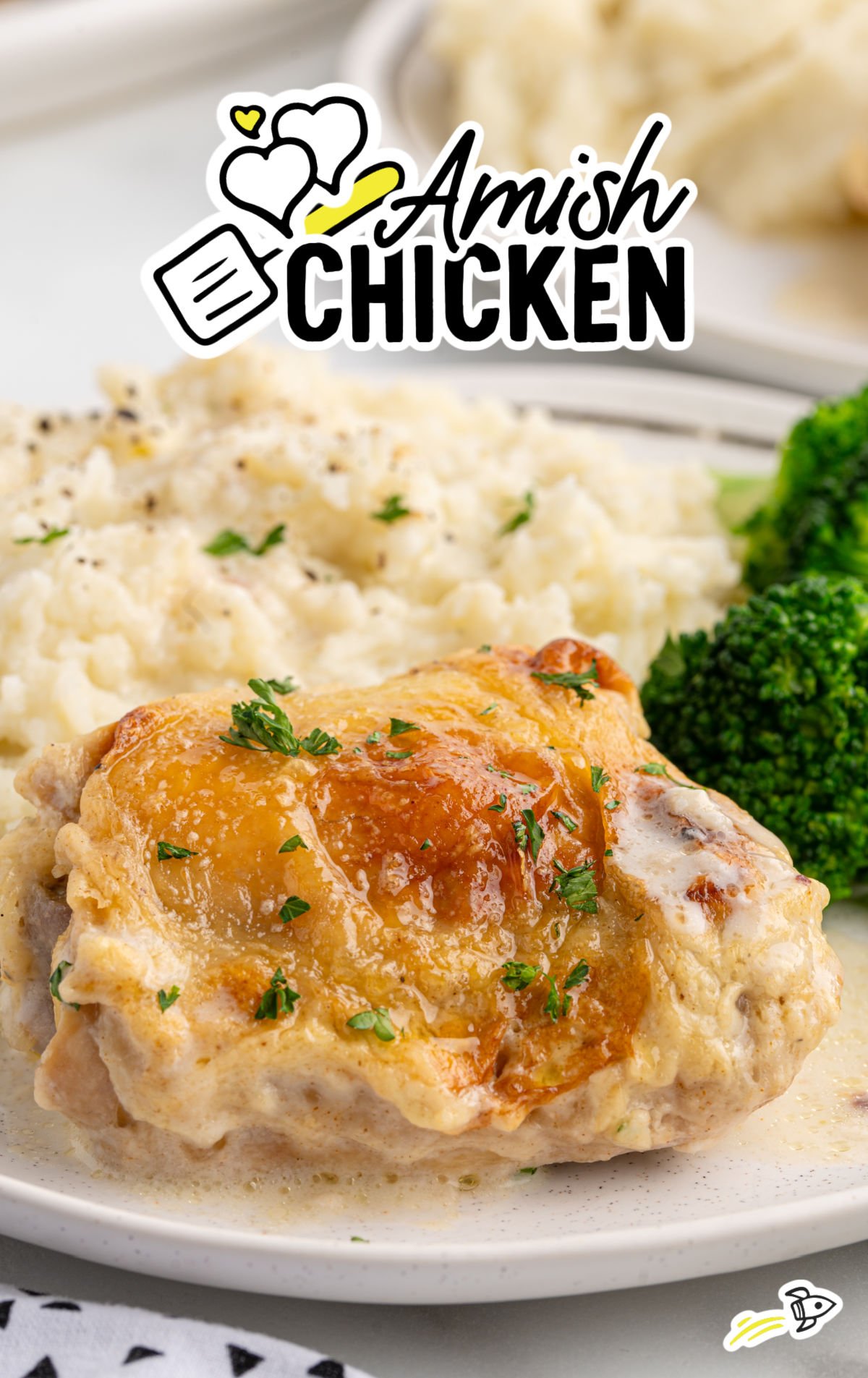 Close-up of a plate featuring golden-brown Amish chicken thigh topped with creamy gravy, served with mashed potatoes and steamed broccoli. The image includes a playful “Amish Chicken” graphic overlay with hearts and a spatula icon.
