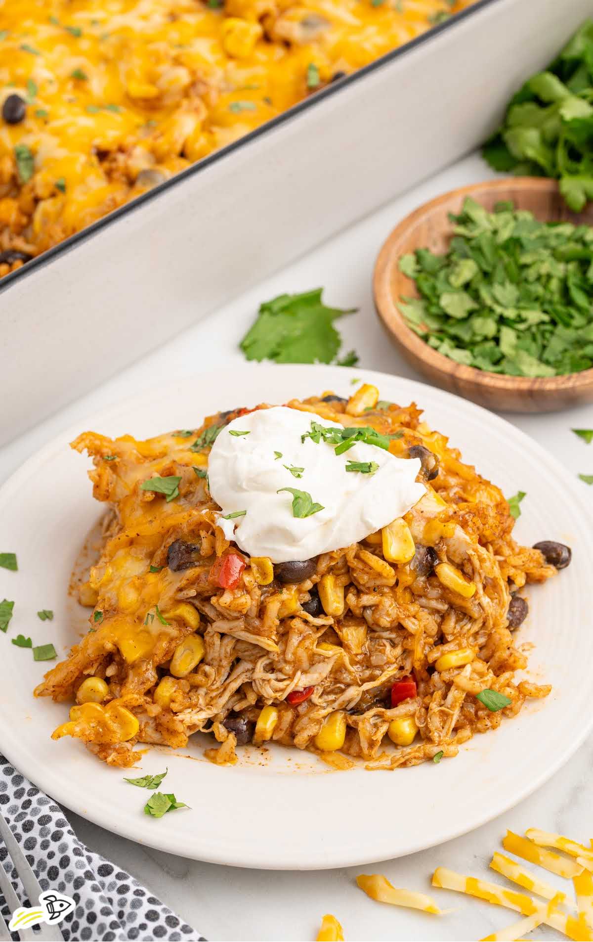 A plate with a generous serving of chicken enchilada rice casserole topped with a dollop of sour cream and fresh cilantro, sitting next to bowls of garnish and a casserole dish.