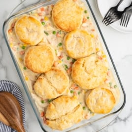 A glass baking dish filled with creamy Chicken Pot Pie Casserole, topped with eight golden-brown biscuits. A wooden spoon, striped napkin, and three stacked plates with forks are nearby on a marble surface.
