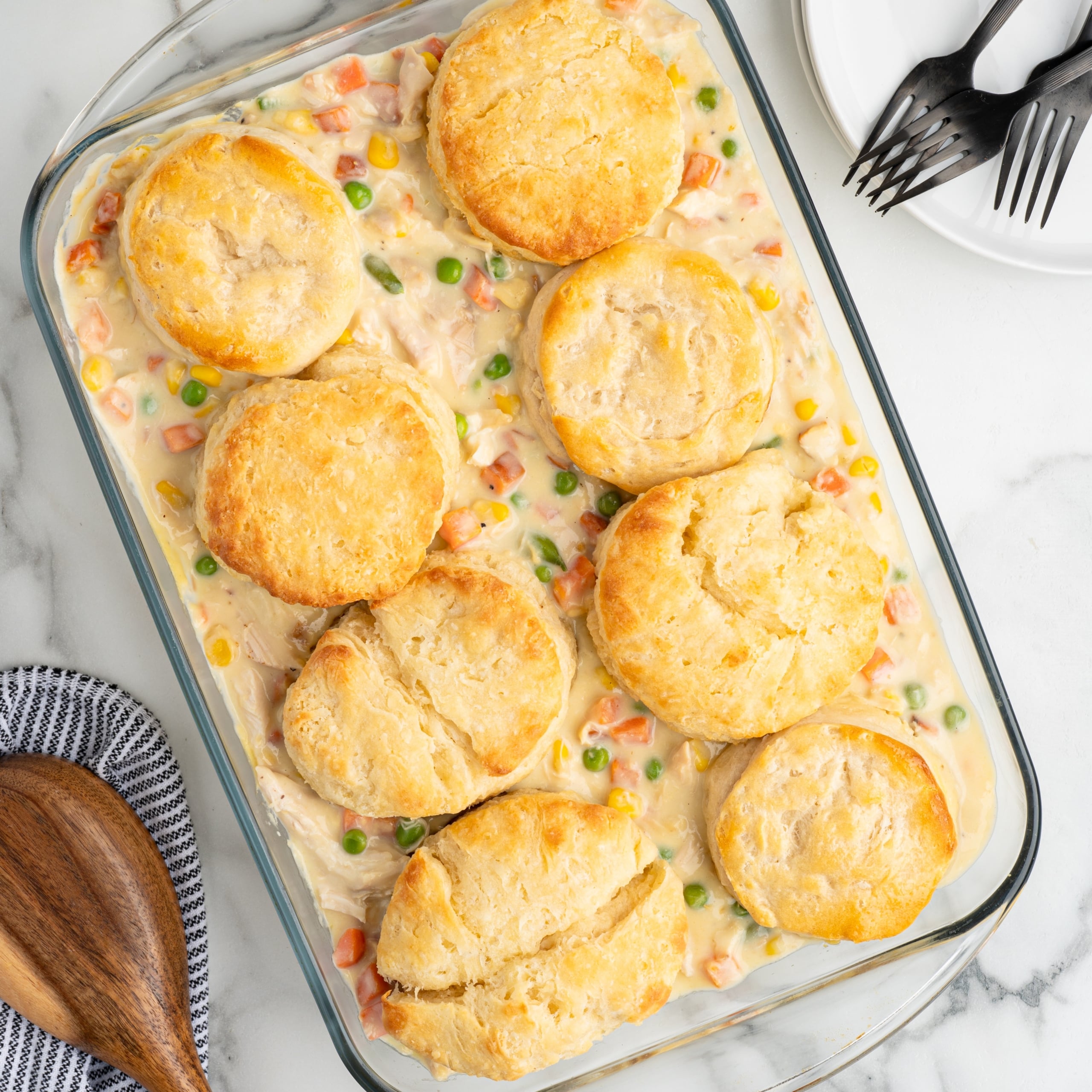 A glass baking dish filled with creamy Chicken Pot Pie Casserole, topped with eight golden-brown biscuits. A wooden spoon, striped napkin, and three stacked plates with forks are nearby on a marble surface.