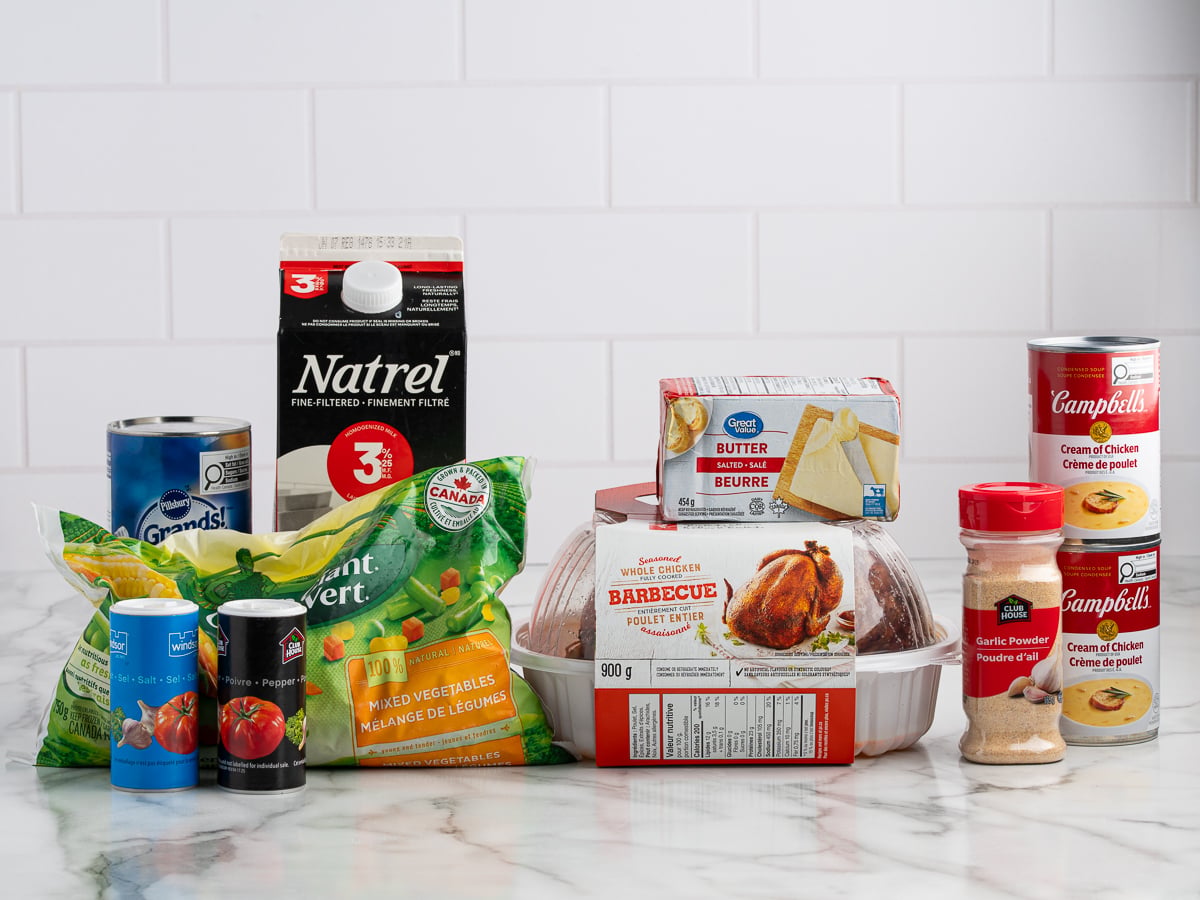 A variety of groceries on a marble surface, including milk, canned soup, frozen vegetables, butter, rotisserie chicken for Chicken Pot Pie Casserole, biscuit dough, canned tomatoes, and a seasoning jar—all against a white tiled background.