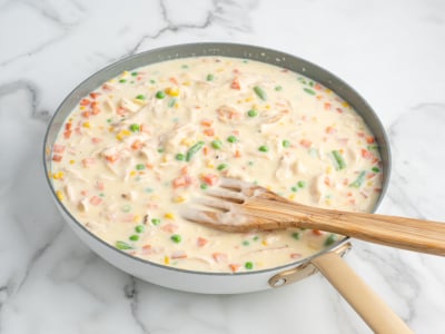 A skillet filled with creamy mixed vegetable sauce, perfect for a Chicken Pot Pie Casserole, including peas, carrots, green beans, and corn, being stirred with a wooden spatula on a white marble surface.