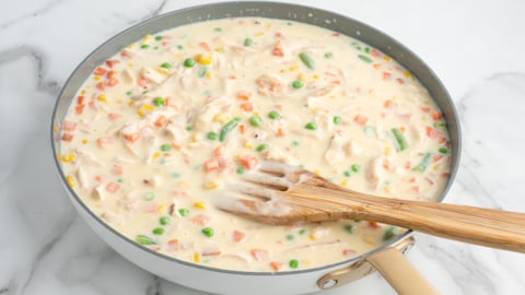 A skillet filled with creamy mixed vegetable sauce, perfect for a Chicken Pot Pie Casserole, including peas, carrots, green beans, and corn, being stirred with a wooden spatula on a white marble surface.