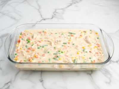 A glass baking dish filled with an uncooked Chicken Pot Pie Casserole mixture—creamy sauce, diced carrots, green beans, corn, peas, and chunks of chicken—sits on a white marble surface.