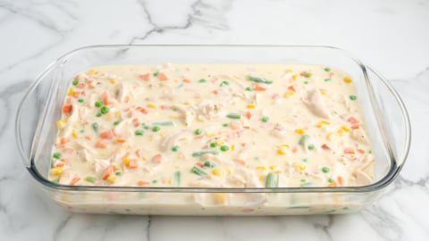 A glass baking dish filled with an uncooked Chicken Pot Pie Casserole mixture—creamy sauce, diced carrots, green beans, corn, peas, and chunks of chicken—sits on a white marble surface.